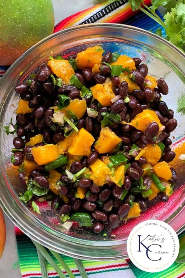 Close up of a glass bowl filled with black bean mango salad with logo on right corner.