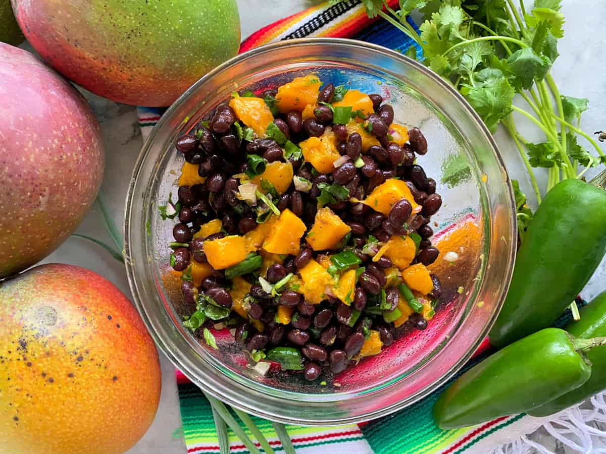 Top view of a bowl filled with black beans and mangoes on top of a kitchen towel with fresh jalapenos and manges.