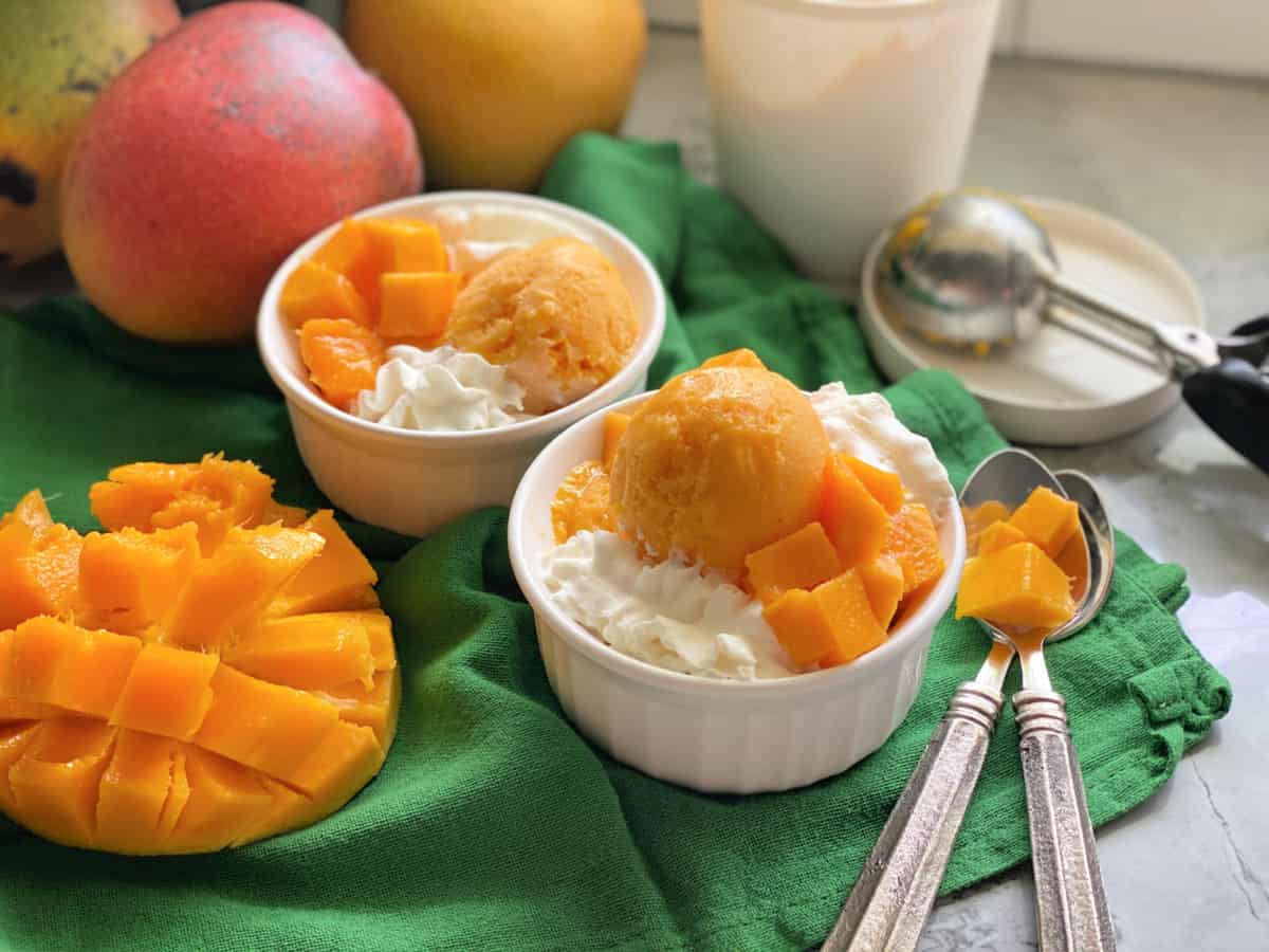 Two white bowls filled with Mango Frozen Yogurt, and a diced mango on a green cloth with spoons.