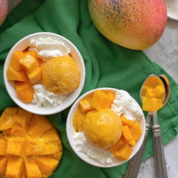 Top view of a two white bowls filled with orange ice cream with whipped cream and mangoes on the counter.