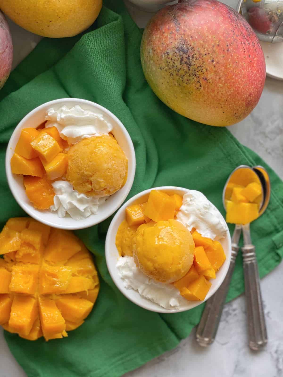 Top view of a two white bowls filled with orange ice cream with whipped cream and mangoes on the counter.