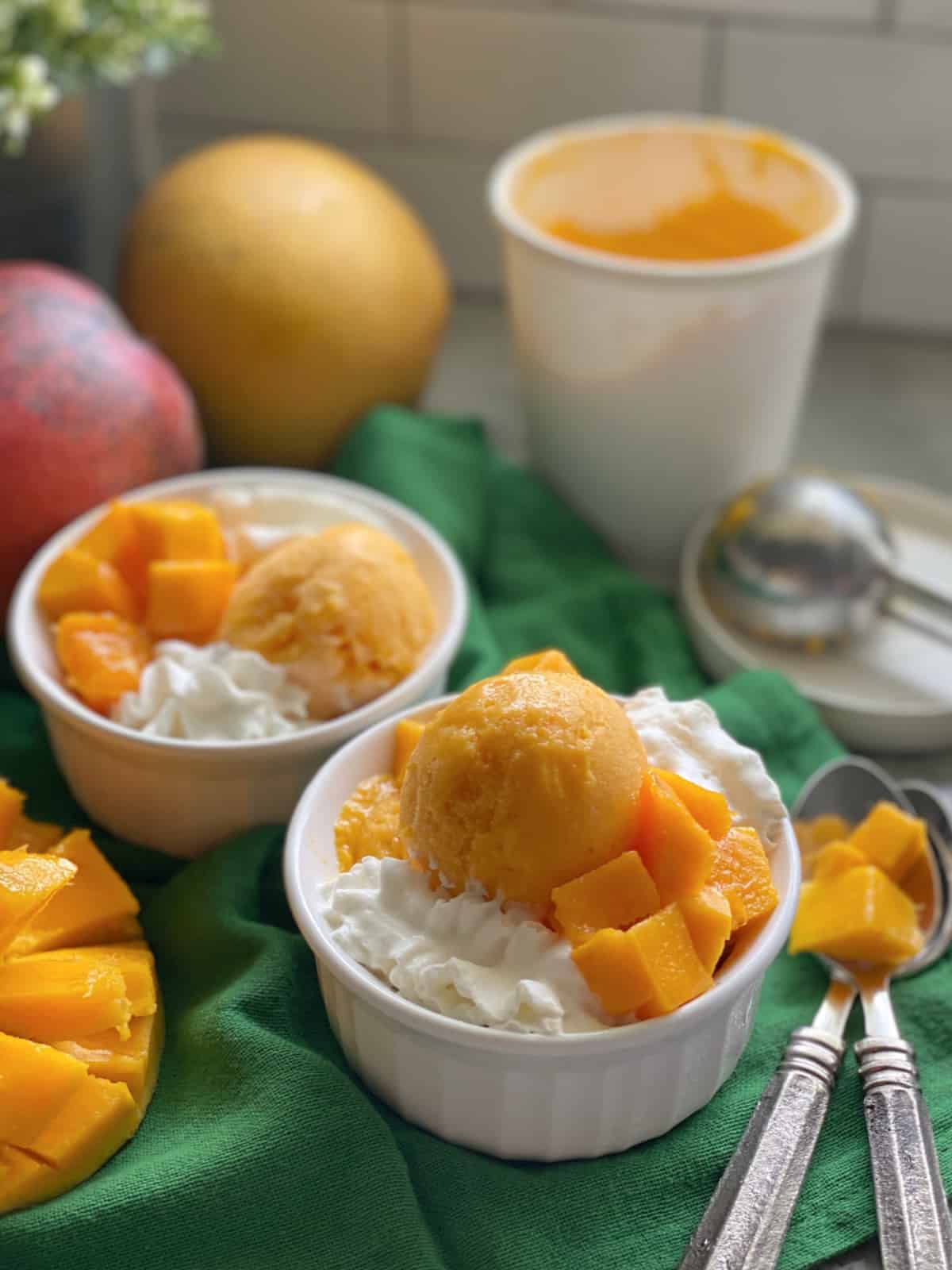 Two white bowls filled with Mango Frozen Yogurt on a green cloth with spoons and mangoes on the counter.