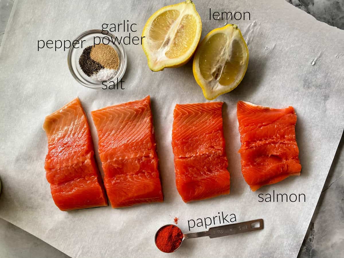 Ingredients on white parchment paper: salmon, paprika, salt, pepper, garlic powder, and lemon.