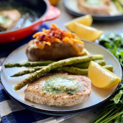White plate filled with a swordfish steak with butter, asparagus, and a baked potato.
