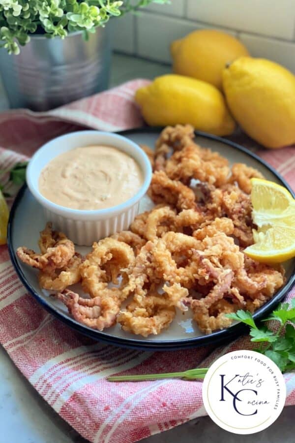 White plate filled with fried squid with lemon wedge and sauce in white ramekin with logo on right corner.