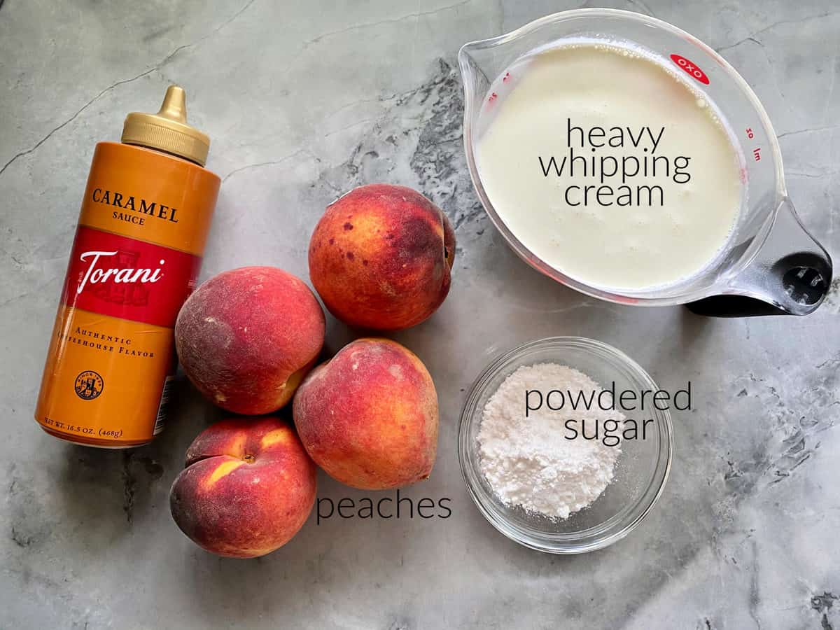 Ingredients on marble countertop: caramel sauce, peaches, whipping cream in measuring cup, and powdered sugar.