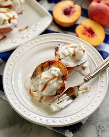 Two grilled peaches with whipped cream and caramel sauce on a white plate with a fork covered in whipped cream.