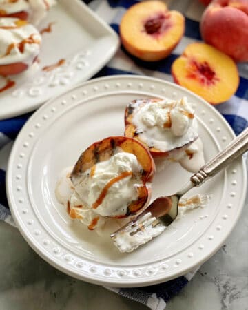 Two grilled peaches with whipped cream and caramel sauce on a white plate with a fork covered in whipped cream.