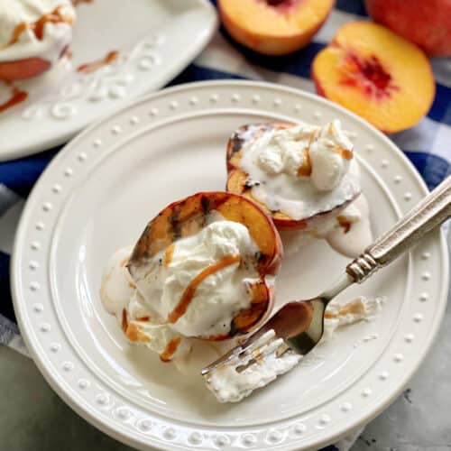 Two grilled peaches with whipped cream and caramel sauce on a white plate with a fork covered in whipped cream.