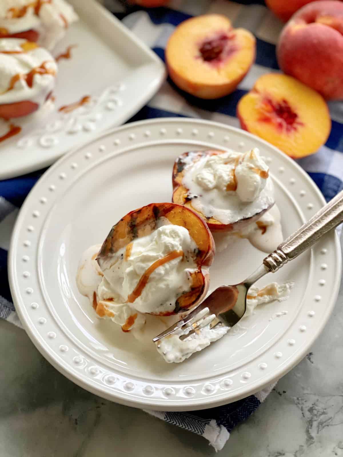 Two grilled peaches with whipped cream and caramel sauce on a white plate with a fork covered in whipped cream.