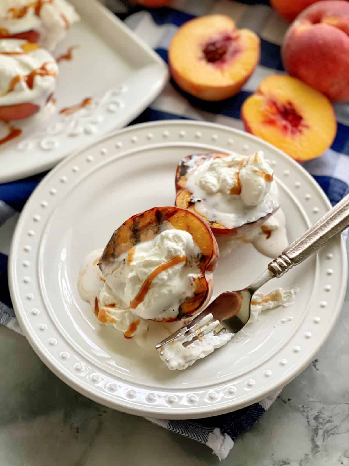 Grilled Peaches with Caramel Sauce and Whipped Cream Two grilled peaches with whipped cream and caramel sauce on a white plate with a fork covered in whipped cream.