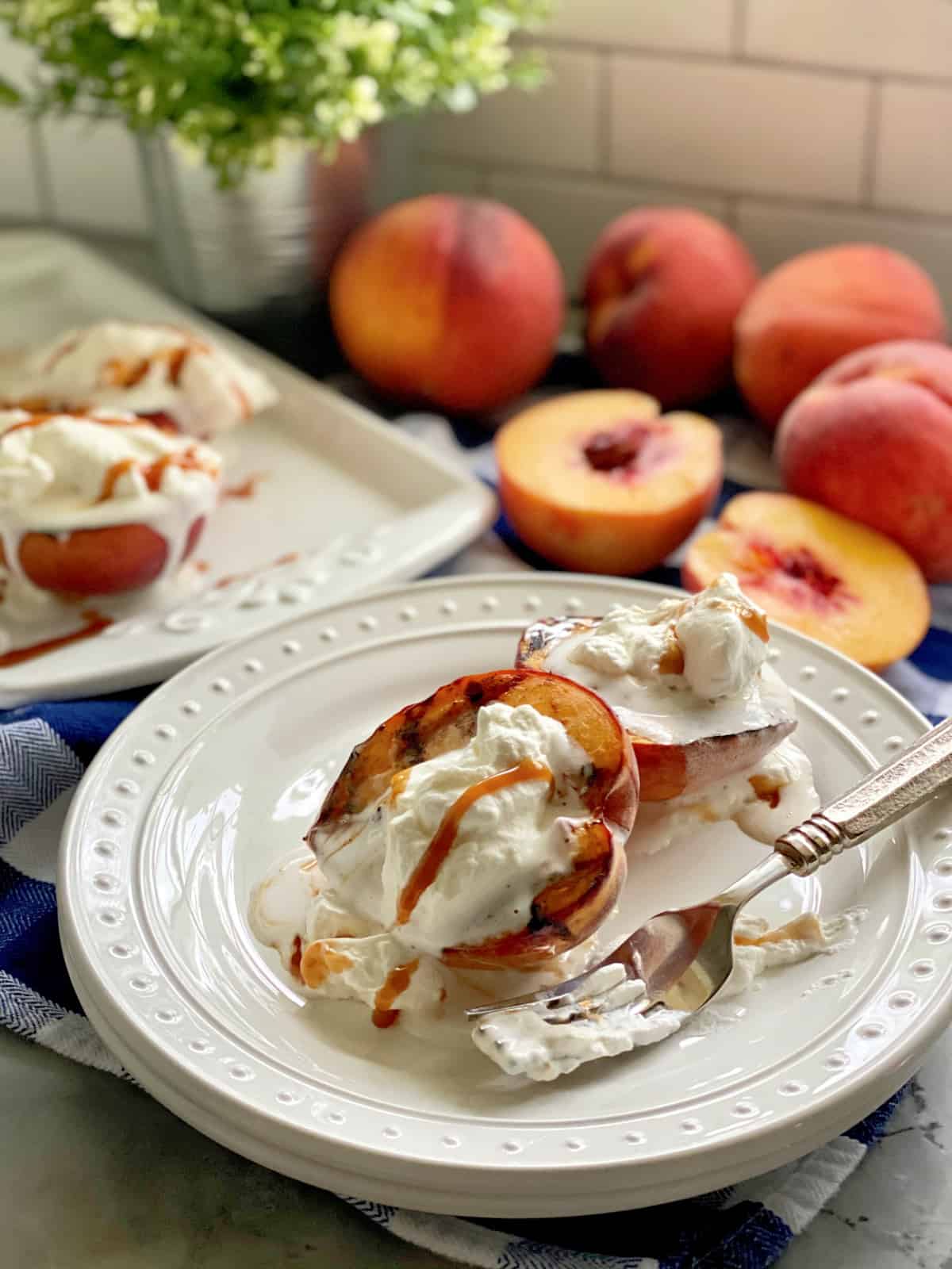 Grilled Peaches with Caramel Sauce and Whipped Cream Two white plates stacked topped with grilled peaches and cream with peaches in background.