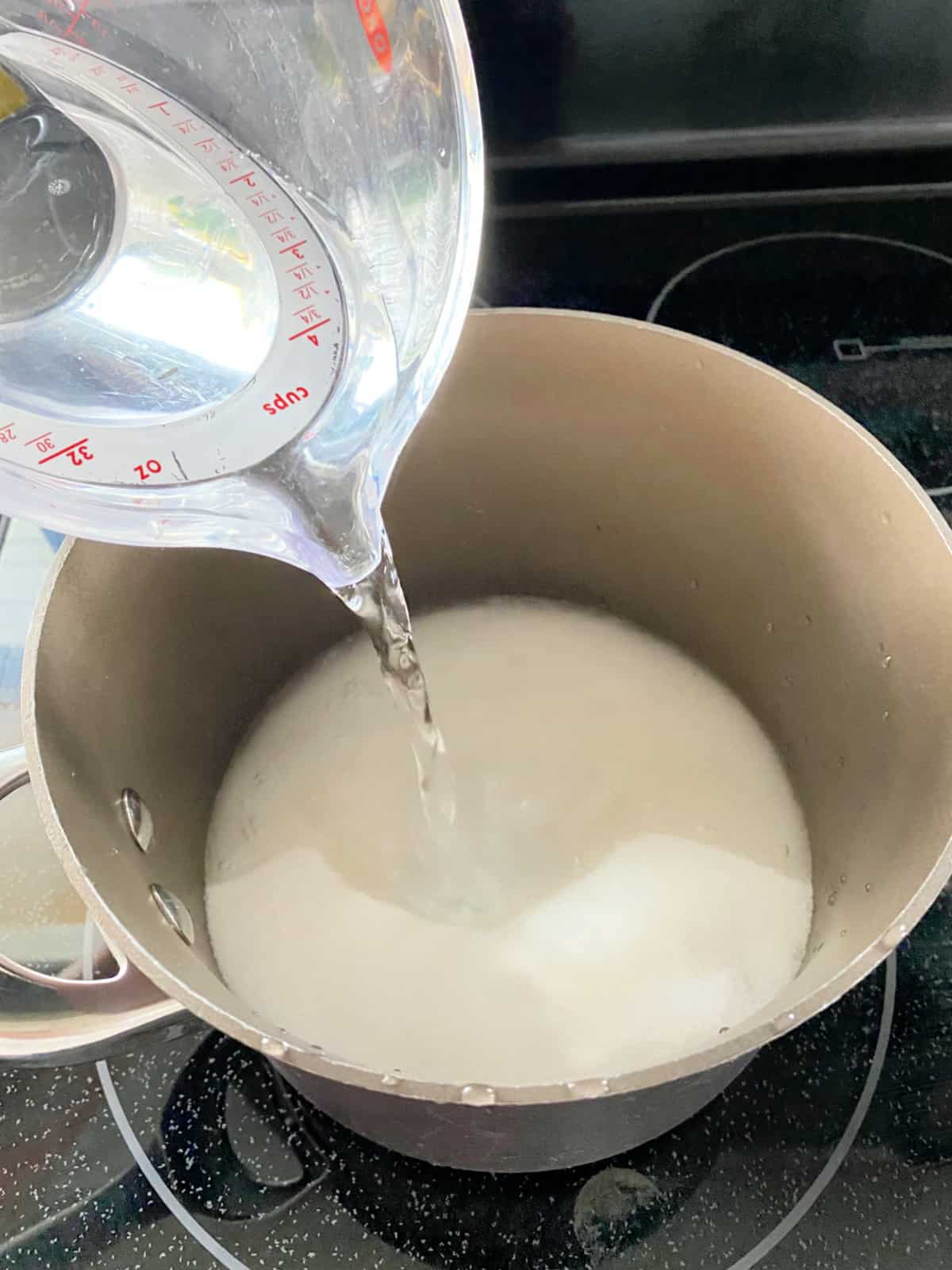 Measuring cup pouring water into a pot filled with sugar.