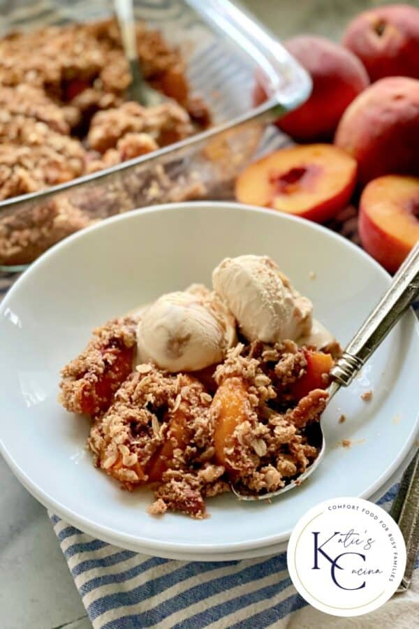 White shallow bowl filled with peach crisp, ice creama, and a spoon with logo on right corner.