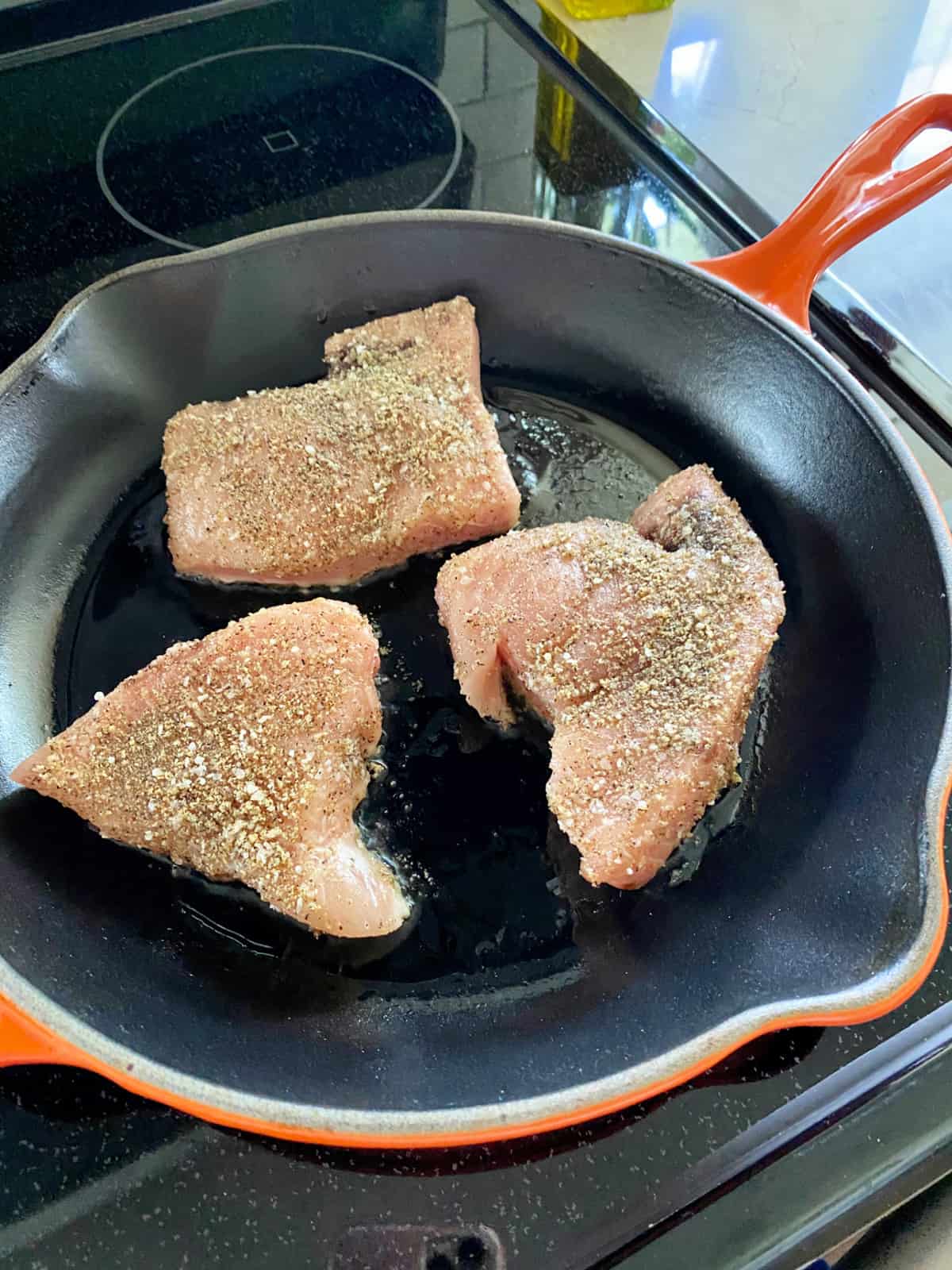 Orange cast iron skillet on a glass stove top with 3 seasoned Swordfish Steaks in the skillet.