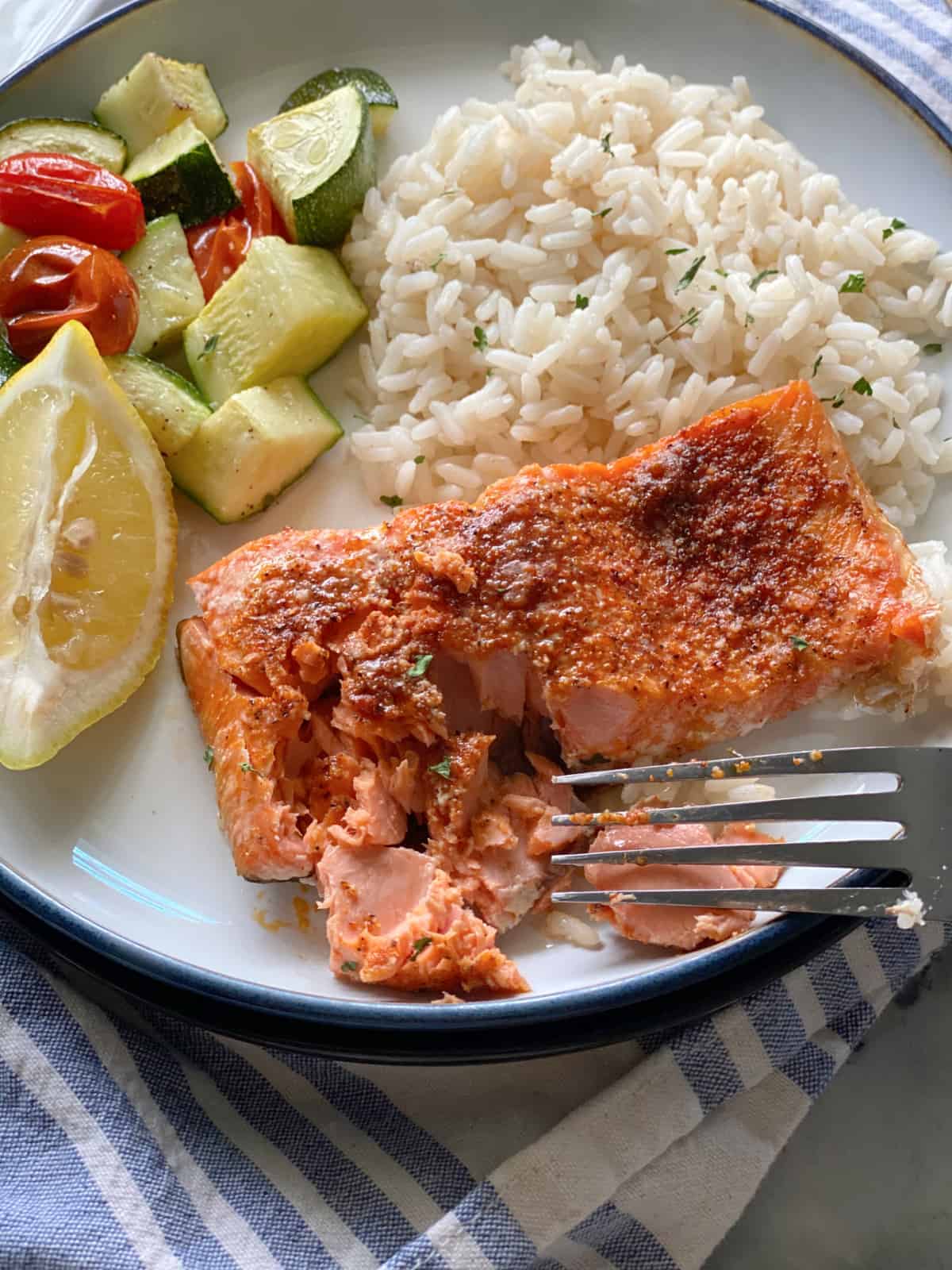 White plate filled with white rice, zucchini, tomatoes, a lemon wedge and a fork flaking cooking salmon.