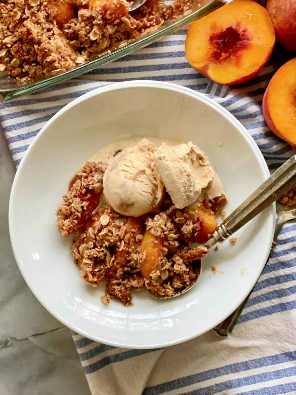 Top view of peach crisp with ice cream in a shallow white bowl filled with fresh peaches around it.
