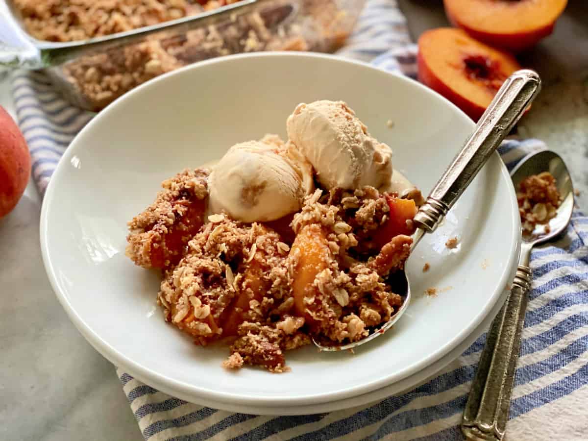 White bowl filled with peach crisp with ice cream and a silver spoon.