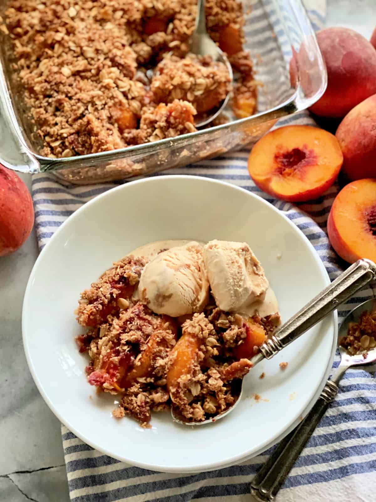 Top view of a white bowl filled with peach crisp, ice cream, a spoon with a glass baking dish filled with peach crisp next to it.