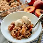 White shallow bowl filled with peach crisp and caramel ice cream with a spoon and peaches in the background.