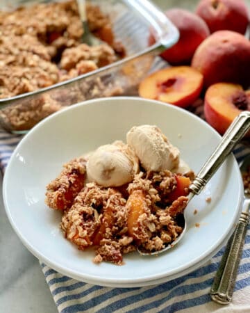 White shallow bowl filled with peach crisp and caramel ice cream with a spoon and peaches in the background.