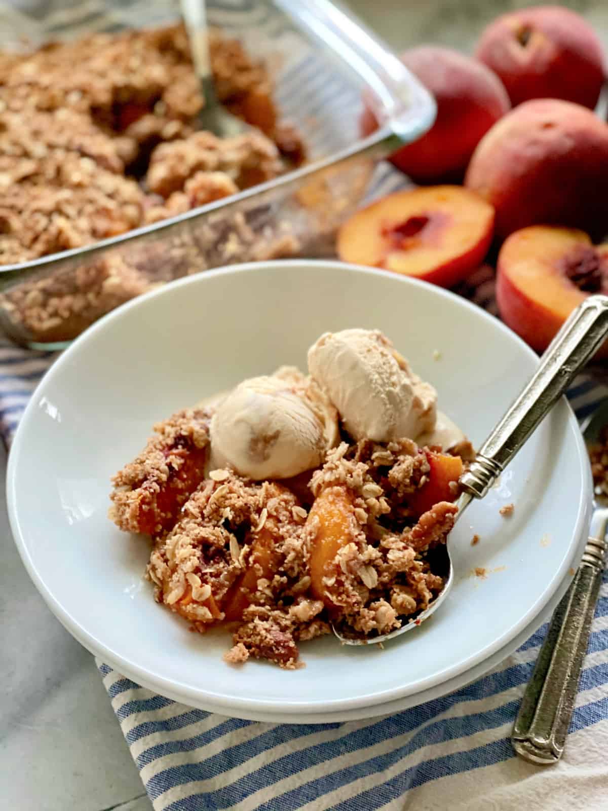 White shallow bowl filled with peach crisp and caramel ice cream with a spoon and peaches in the background.