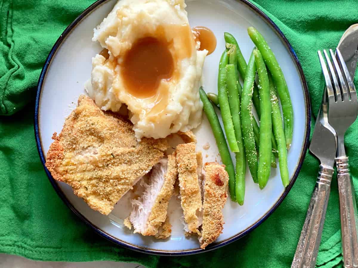 Top view of a white plate with sliced pork chop, mashed potato with gravy, and green beans.