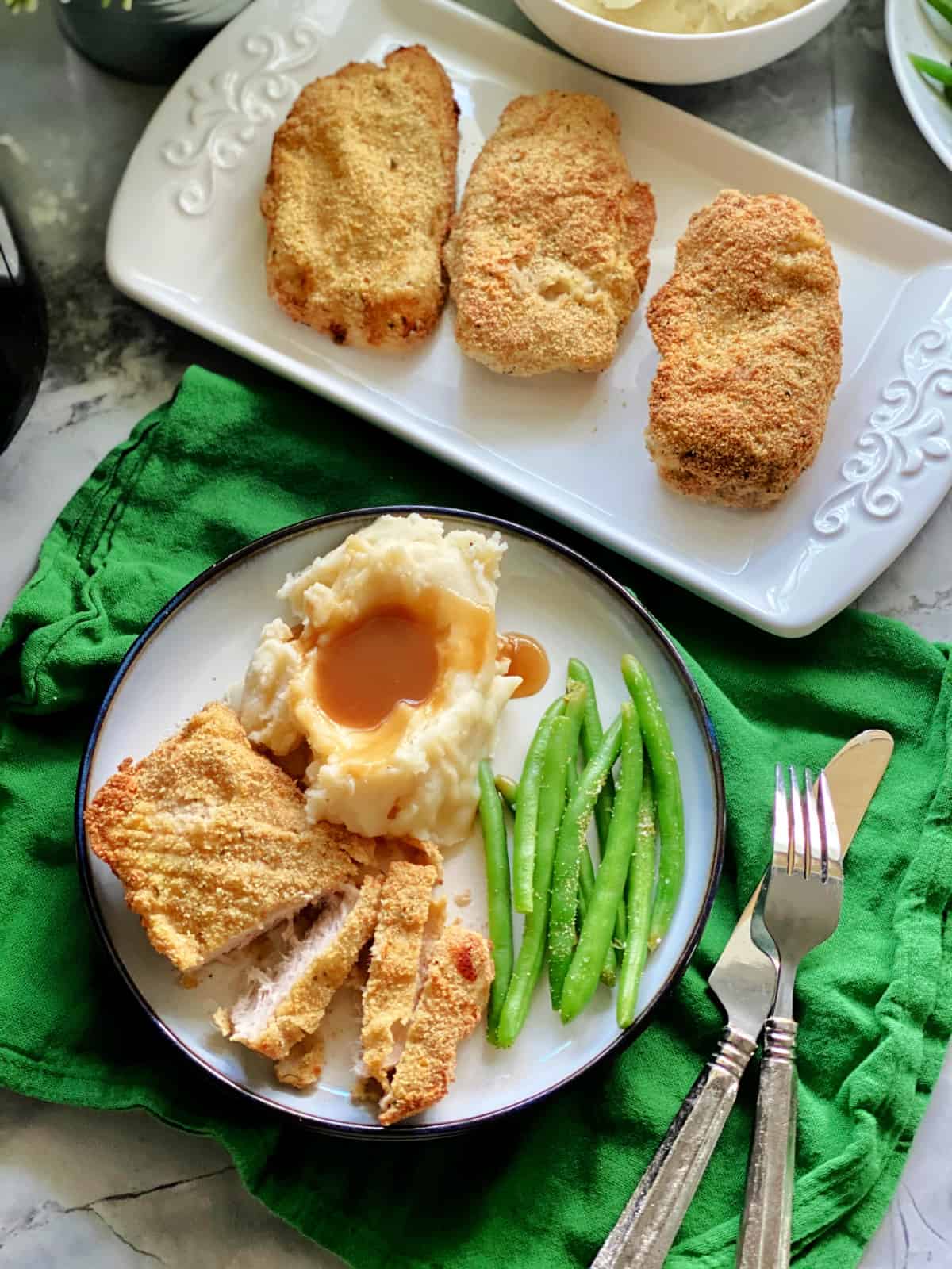 Top view of a white plate filled with pork chop slices, mashed potatoes, and green beans with pork chops on a platter next to it.