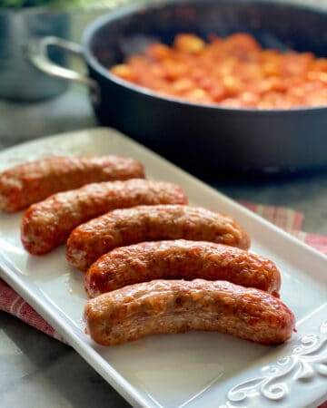 White platter filled with crispy sausage links with tomato red sauce in a skillet in background.