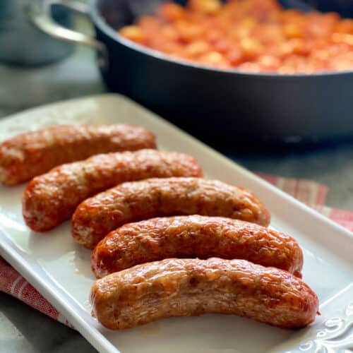 White platter filled with crispy sausage links with tomato red sauce in a skillet in background.
