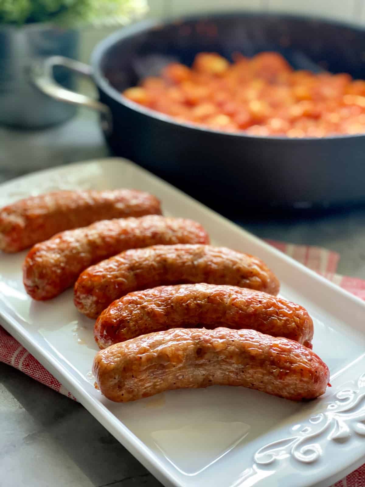 White platter filled with crispy sausage links with tomato red sauce in a skillet in background.