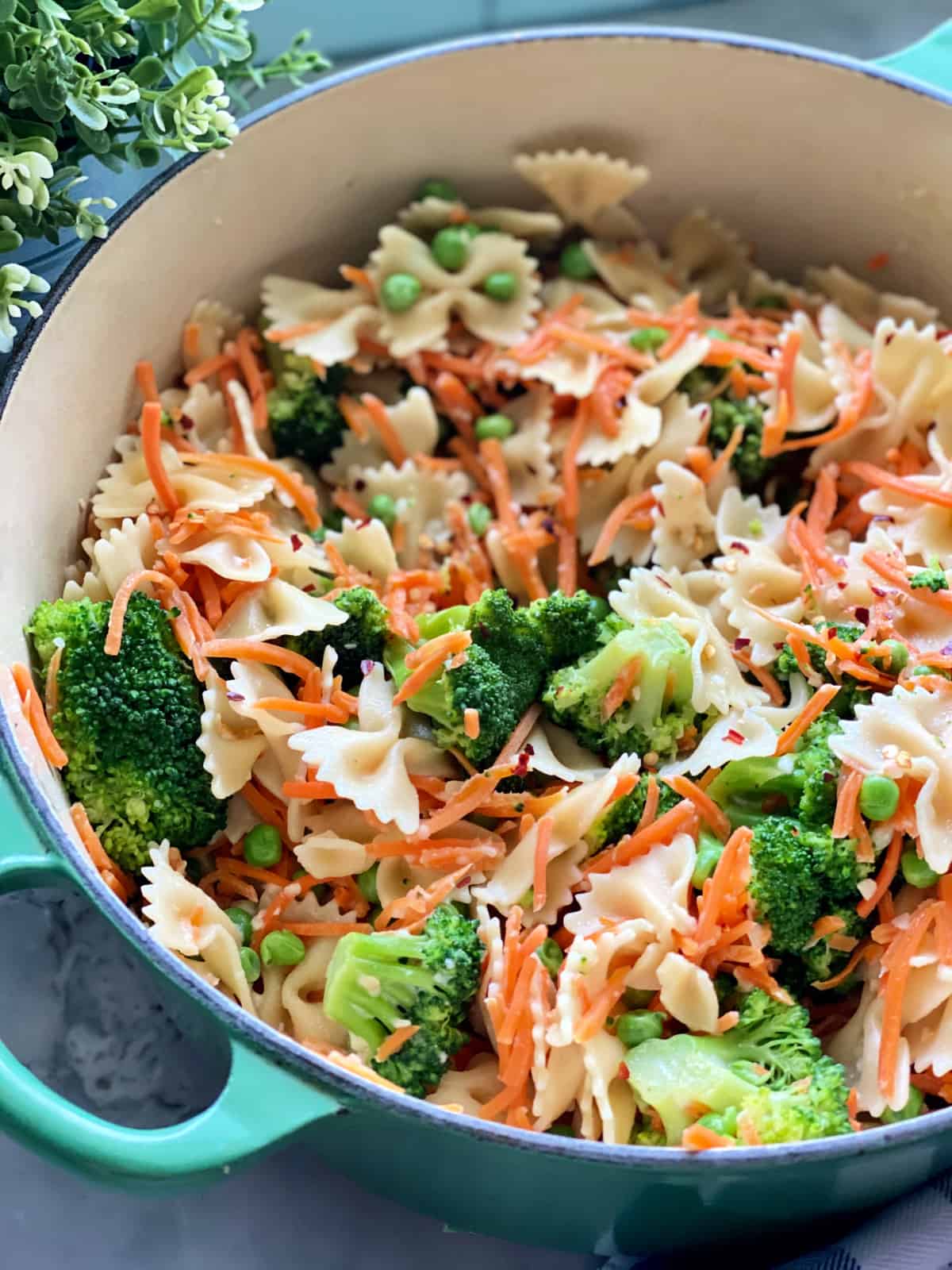 Green pot filled with bow tie pasta, shredded carrots, broccoli, green peas, and red pepper flakes. 