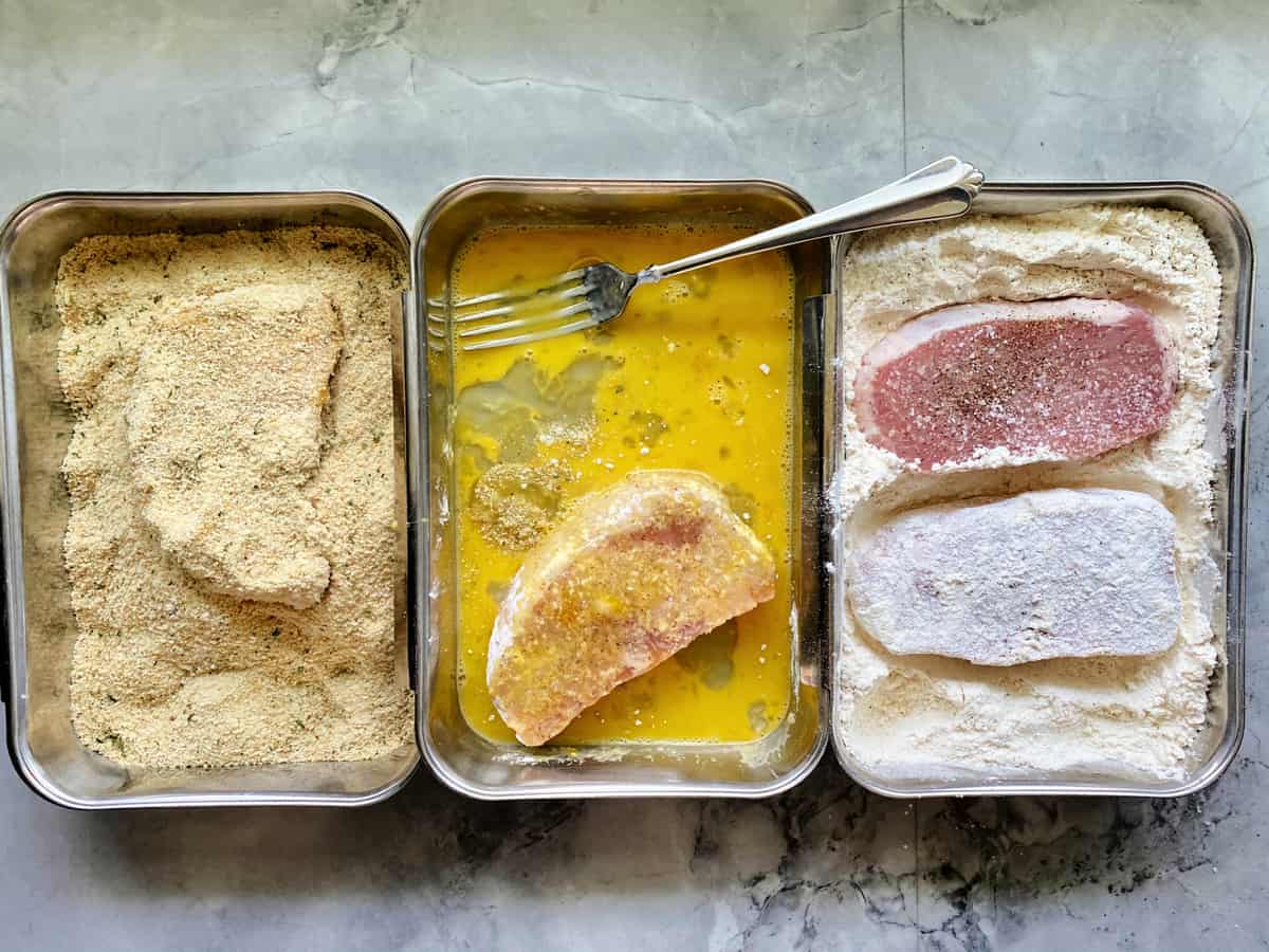 Three trays with the process of breading pork chops; flour, egg wash, and bread crumbs with raw pork chops.