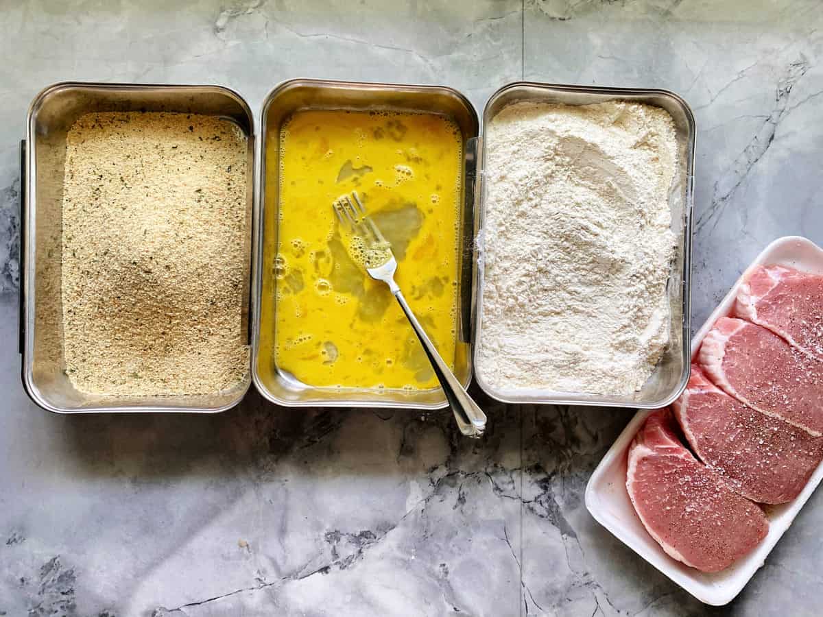 Three trays filled with bread crumbs, egg wash, and flour with raw pork chops in tray next to it.