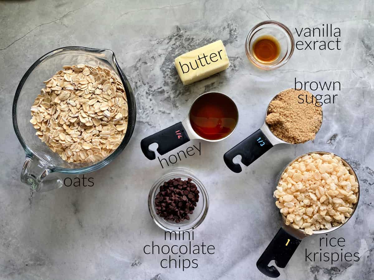 Ingredients on marble countertop: oats, butter, honey, vanila extract, brown sugar, chocolate chips, and rice krispies.