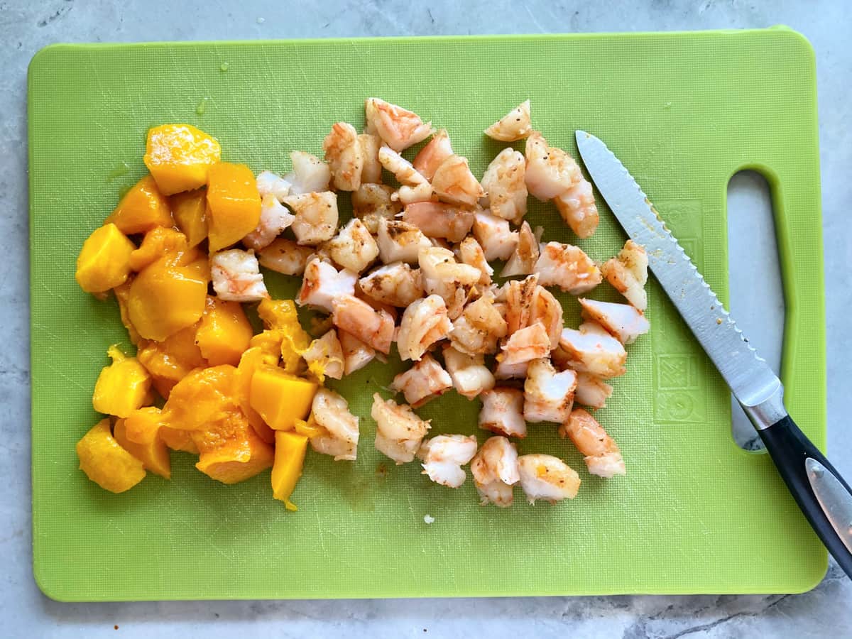 Green cutting board with chopped shrimp and mangos with serrated knife.