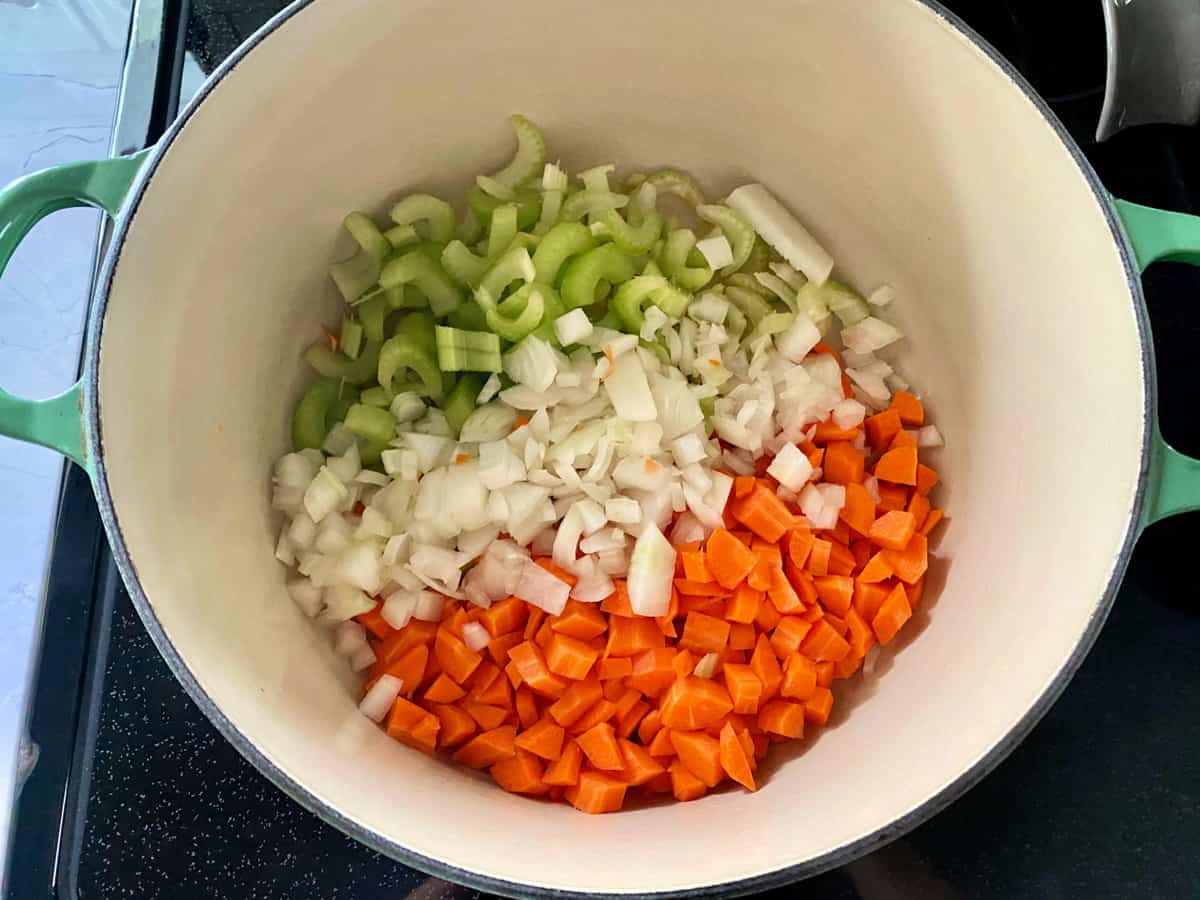 Top view of a white pot filled with diced celery, onions, and carrots on a stovetop.
