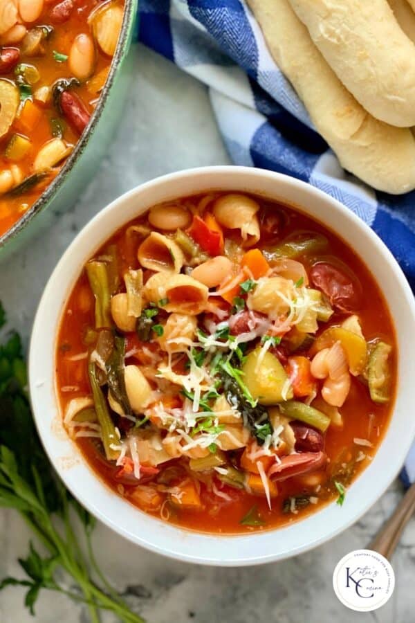 Top view of a white bowl filled with minestrone soup with breadsticks on the side with logo on bottom right corner.