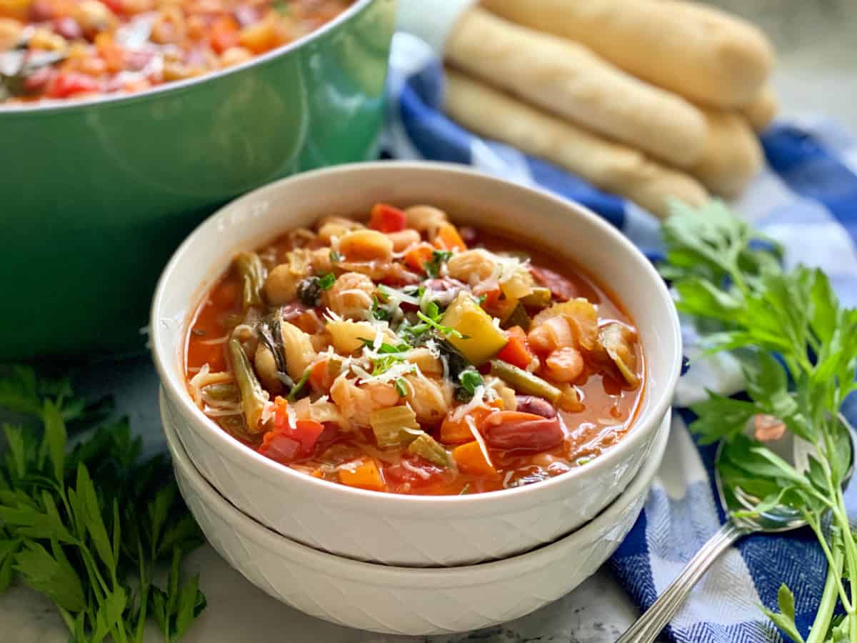 Two white bowls stacked filled with minestrone soup topped with shredded cheese and parsley.