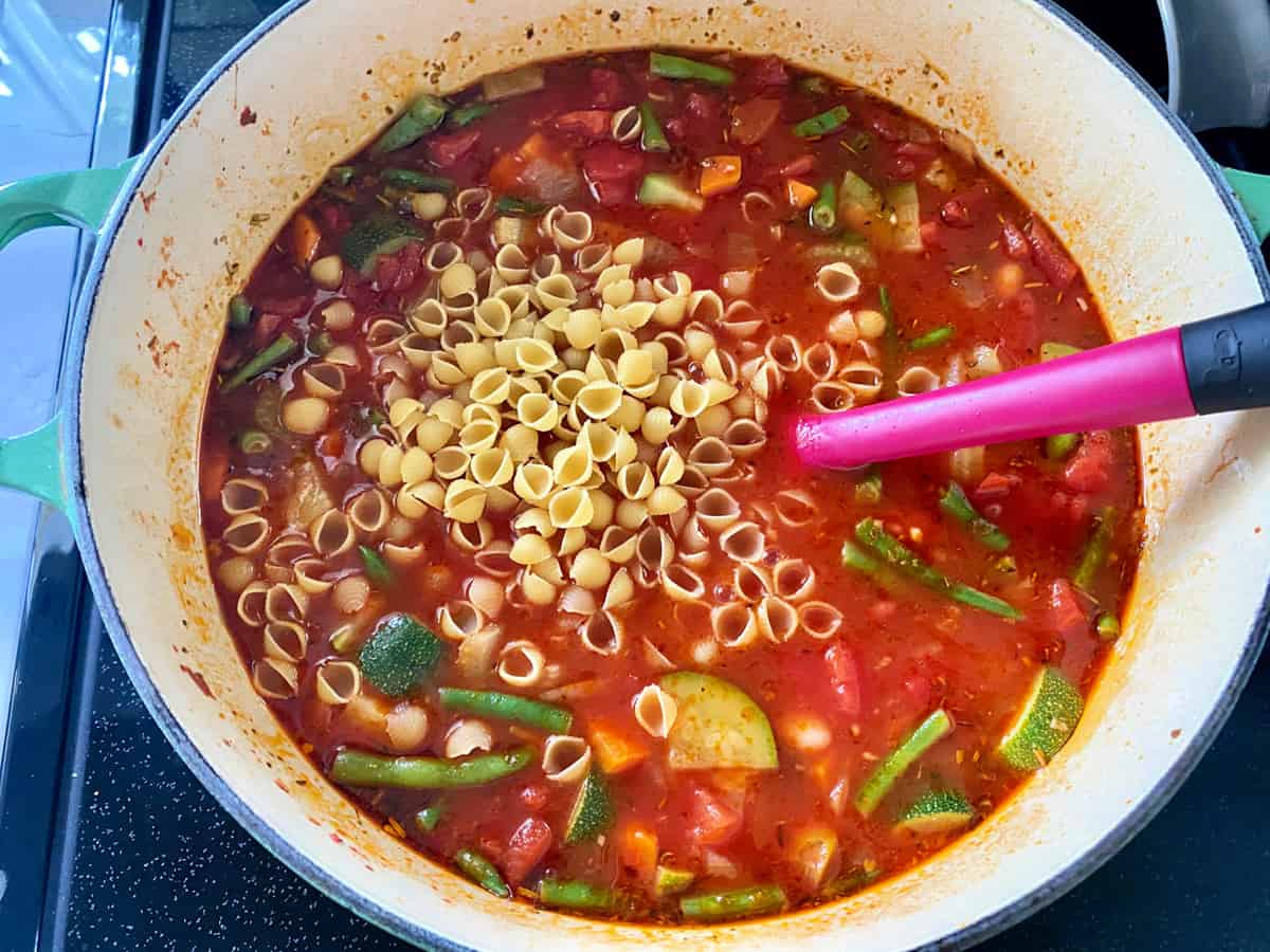 Top view of a white pot filled with red broth, green beans, carrots, zucchini, and shell pasta with a pink spatula.