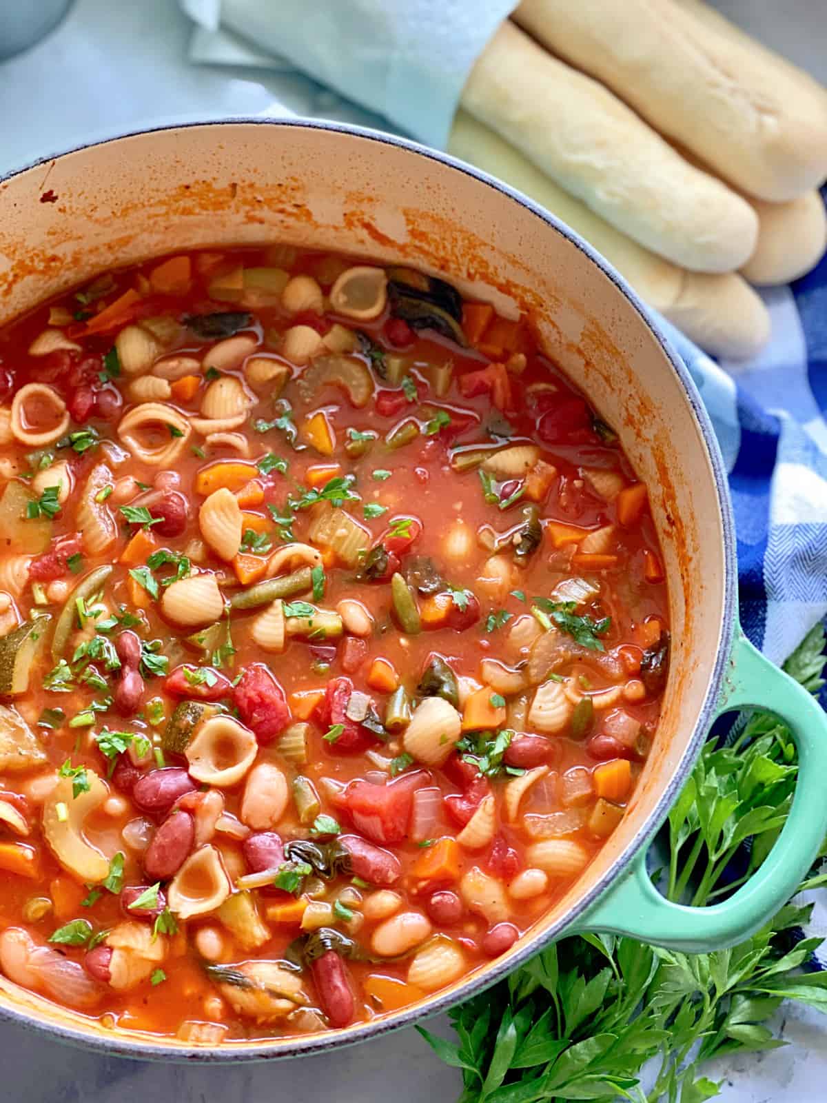 Top view of a big pot of minestrone soup with breadsticks and parsley next to it.