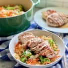 White bowl filled with bow tie pasta, broccoli, carrots, and grilled chicken with grilled chicken in the background and a green pot with pasta in background.