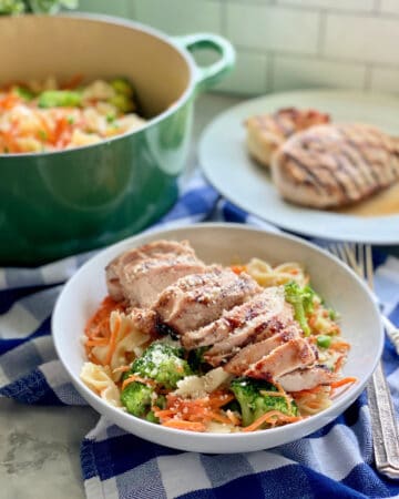 White bowl filled with bow tie pasta, broccoli, carrots, and grilled chicken with grilled chicken in the background and a green pot with pasta in background.