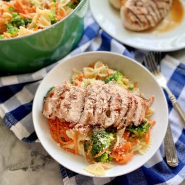 Bowl filled with bow tie pasta with shredded carrots, broccoli, and grilled chicken with a green pot and plate of chicken in the background.
