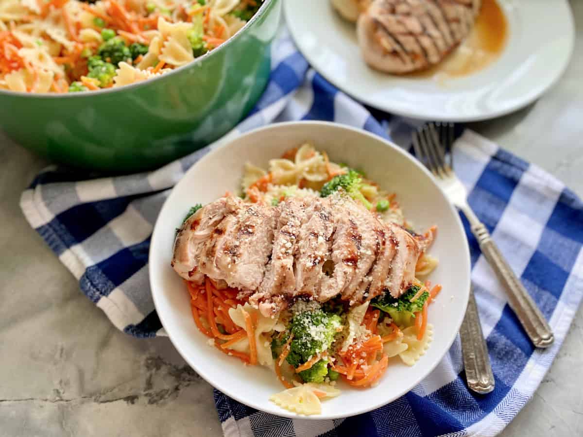 Bowl filled with bow tie pasta with shredded carrots, broccoli, and grilled chicken with a green pot and plate of chicken in the background.