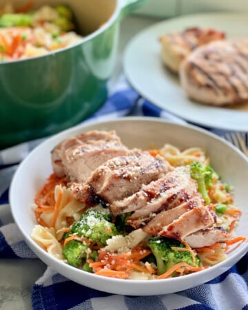 White bowl filled with bow tie pasta, vegetables, and grilled chicken with green pot in background and grilled chicken on plate.