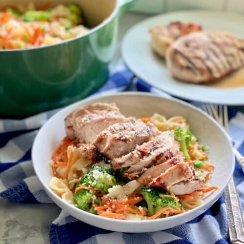 White bowl filled with bow tie pasta, vegetables, and grilled chicken with green pot in background and grilled chicken on plate.