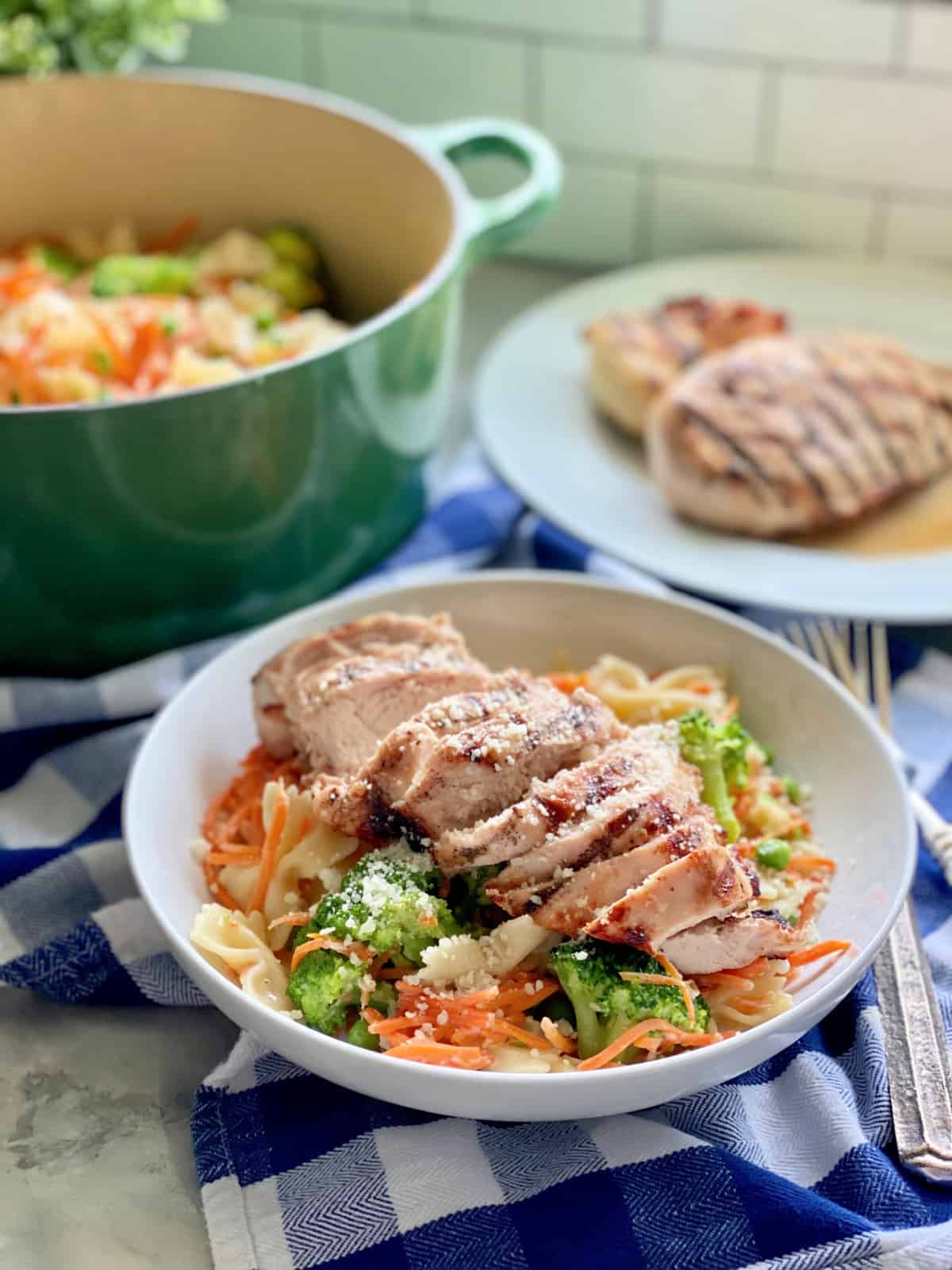White bowl filled with bow tie pasta, broccoli, carrots, and grilled chicken with grilled chicken in the background and a green pot with pasta in background.