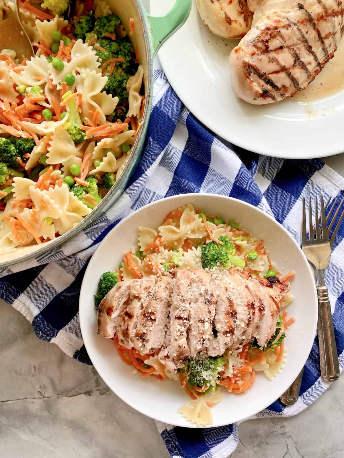 Top view of a bowl of Grilled Chicken Pasta with vegetables, a large pot with bow tie pasta and veggies and a plate of grilled chicken breast.