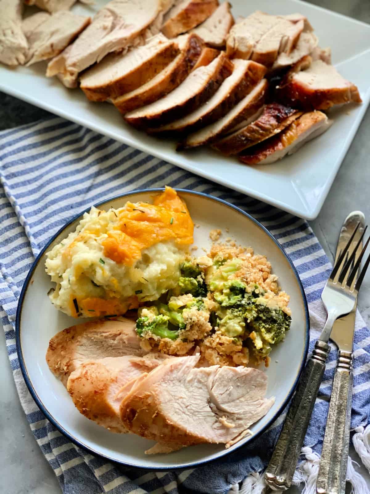 Top view of sliced turkey breast, mashed potatoes, and broccoli casserole with silverware on the side and platter of turkey next to the plate.